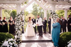 Wedding ceremony taking place under a white gazebo decorated with elegant white floral arrangements.