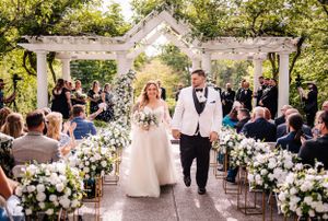 Couple exits down aisle under white pergola adorned with lush floral arrangements during outdoor ceremony.