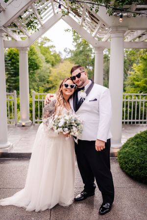 Newlyweds wearing sunglasses pose under a white pergola in an elegant garden setting.