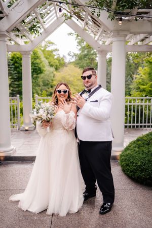 A stylish wedding couple with sunglasses strike a fun pose under a white pergola.