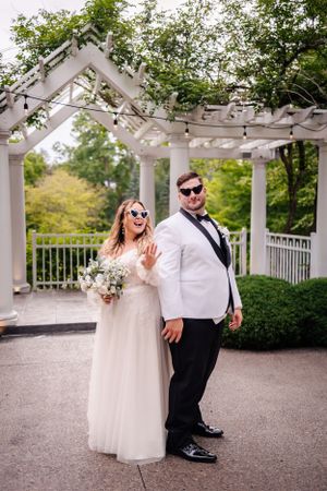 A playful wedding portrait under a decorated pergola with the couple wearing sunglasses.