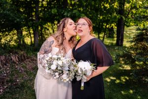 Two celebrants share laughter while holding elegant white floral bouquets in a woodland setting.