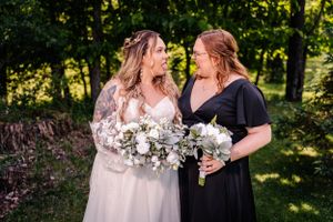 Two people in wedding attire share joyful laughter while holding white flower bouquets outdoors.