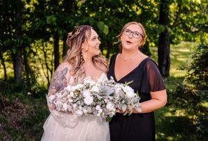 Two people share a candid laughing moment while holding a white floral bouquet in a forest.