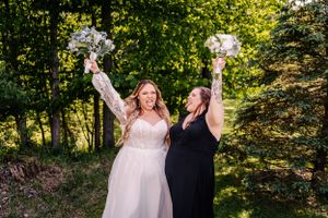 Two celebrants raise their white bouquets triumphantly in a sun-dappled forest setting.