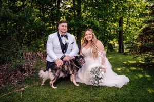 Wedding couple poses with their black and white dog in sunny garden setting.