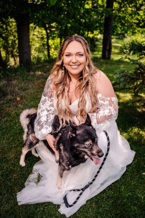A bride in off-shoulder lace gown kneels with black and white dog in wooded garden setting.