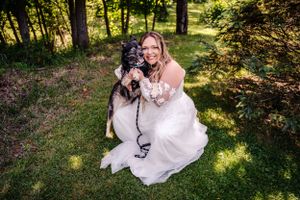 A white wedding dress and a black and white dog pose together in a sunny outdoor woodland setting.