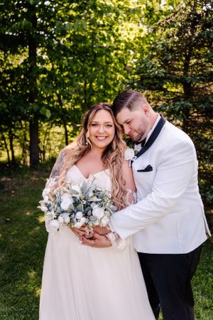 A bride in a flowing dress holds a white and purple bouquet while sharing an intimate moment in a garden setting.