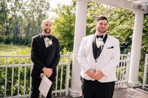 Two groomsmen in formal attire stand on white columned porch during wedding.