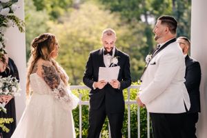 A wedding ceremony takes place under a white columned pergola with guests watching.