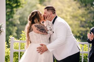 A romantic wedding kiss moment captured under a white pergola surrounded by greenery.