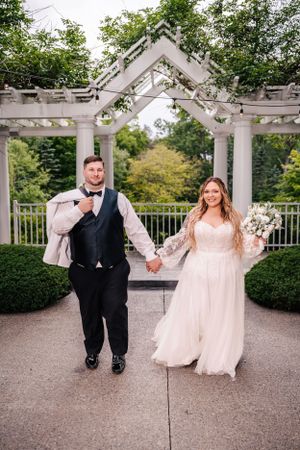 A newlywed couple walks hand-in-hand under an elegant white pergola in a garden setting.