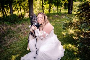 A bride in white lace dress kneels on grass with black and white dog in woodland setting.