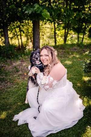 A bride in white gown poses with black and white dog in sunlit forest setting.