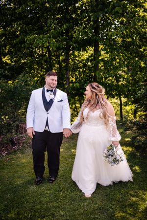 A couple walks hand in hand through a garden, wearing elegant white and black wedding attire.