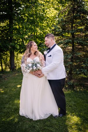 A wedding couple shares a joyful moment in a sunlit garden setting with lush greenery and a beautiful white bouquet.