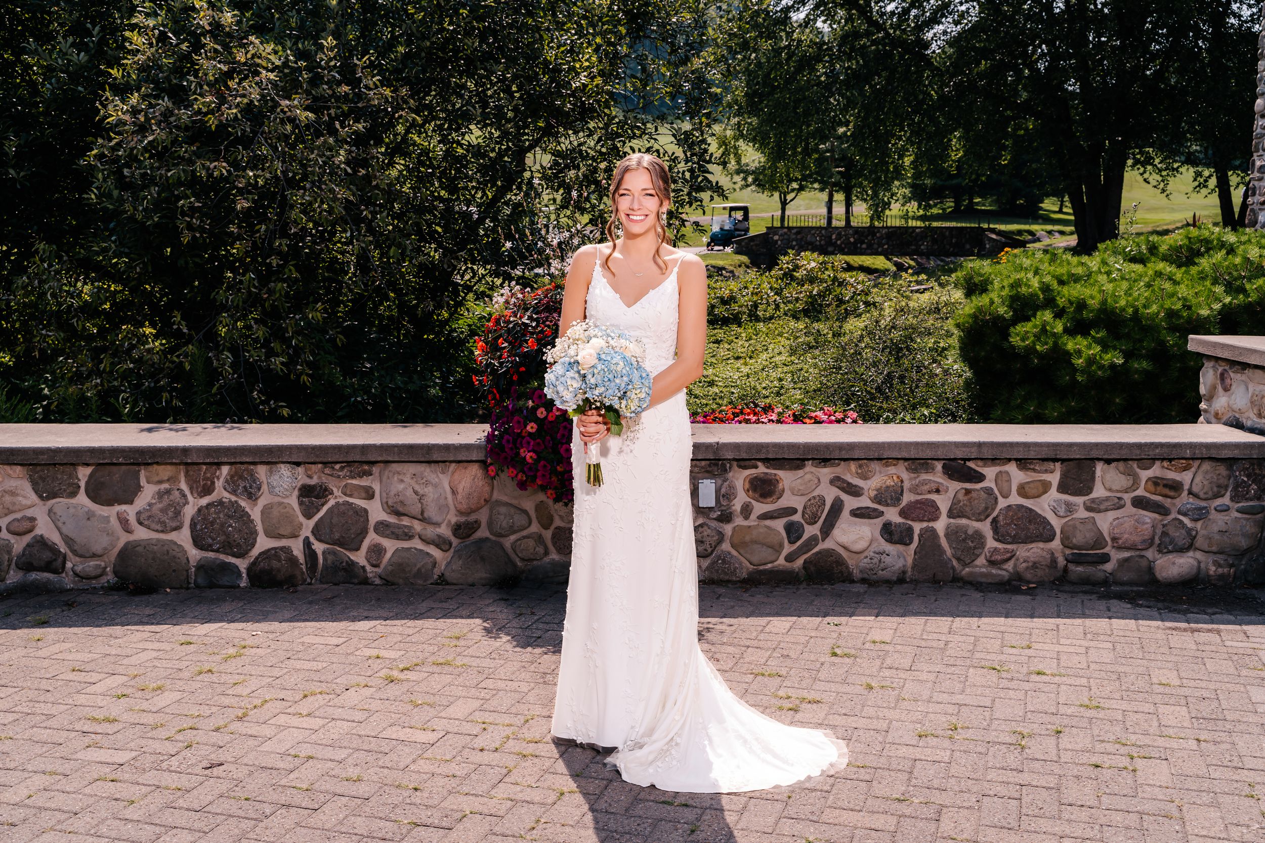 Bride in elegant white gown holds bouquet while posing against stone wall outdoors.