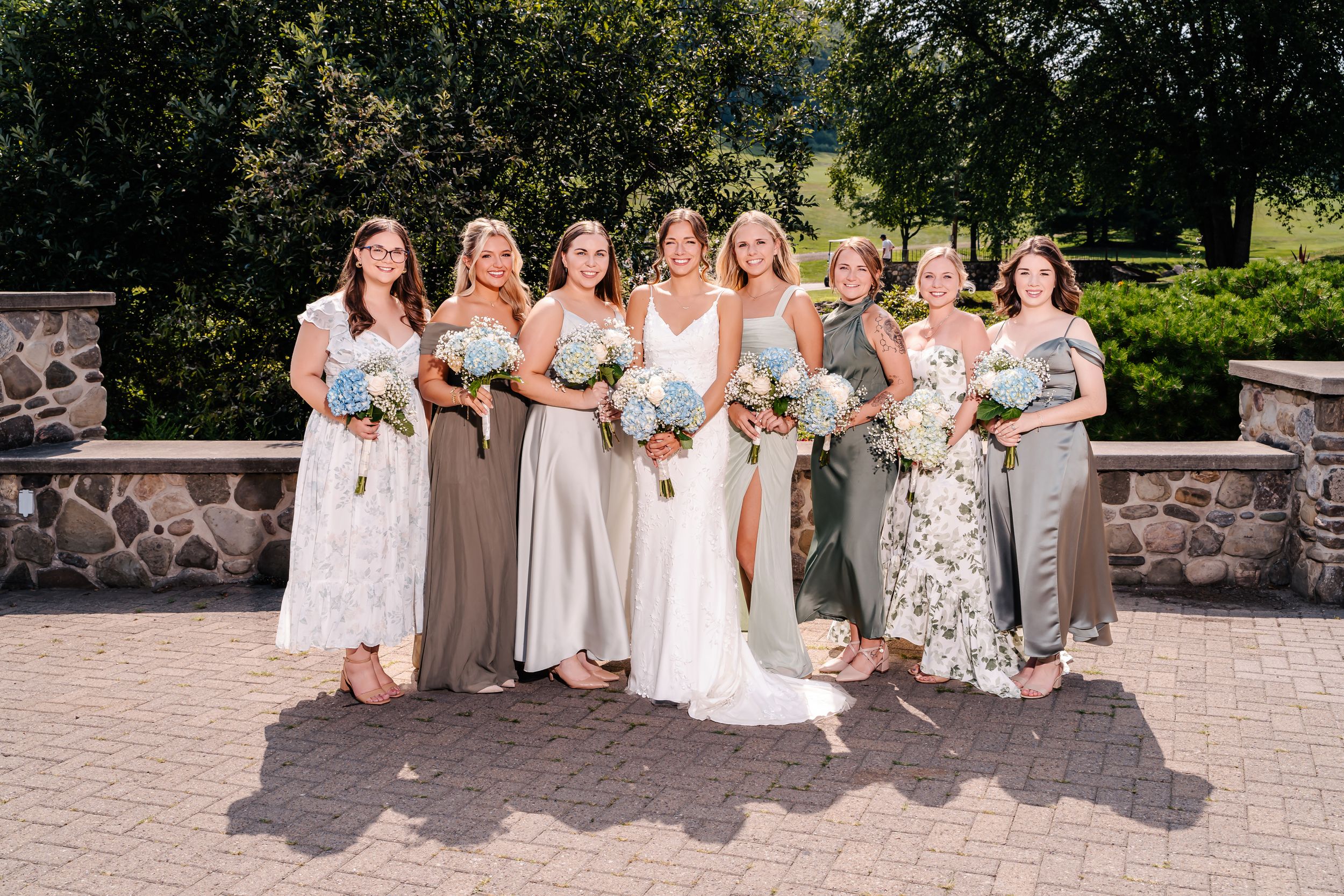Bridal party in mixed floral and solid dresses pose with bouquets on stone patio.