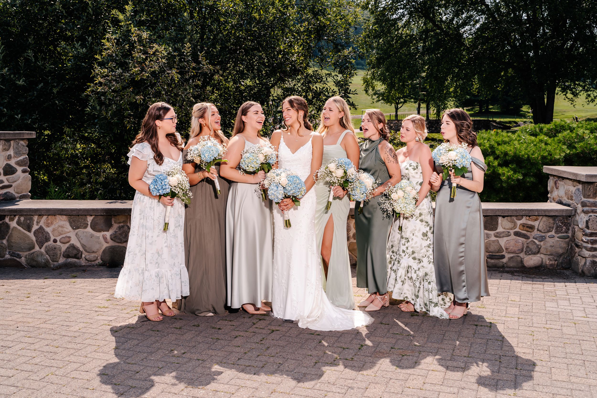 Wedding party of bridesmaids in coordinated dresses pose with bride outdoors.