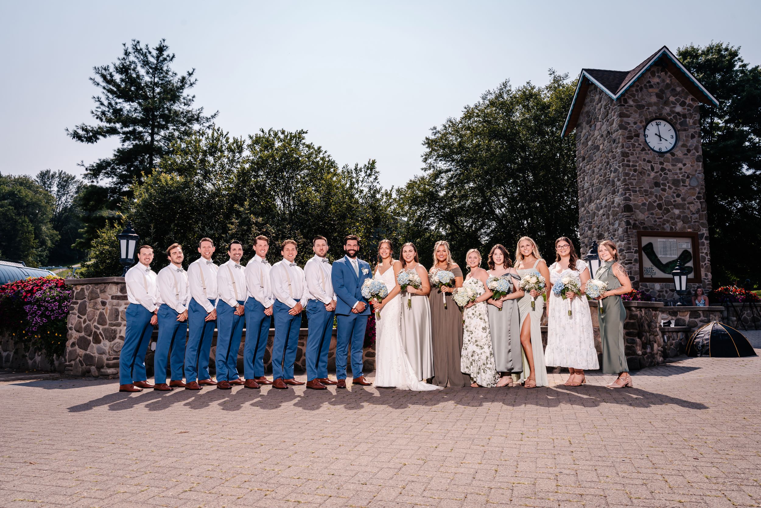 Wedding party lined up outdoors with bridesmaids in floral dresses and groomsmen in blue pants and white shirts.