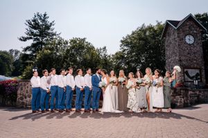 Wedding group photo showing alternating white shirts with blue pants and floral dresses against stone architecture.