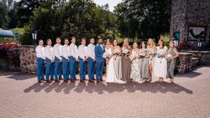 Large wedding party group poses together on brick patio near stone building with clock tower.