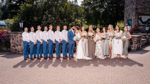 Wedding party members stand together on brick pavers near stone wall and landscaping.
