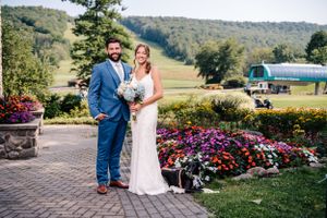 Wedding couple poses in formal attire near colorful flower garden with mountain landscape behind them.