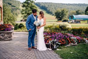 Newlyweds stand together on brick pathway surrounded by vibrant purple flowers and mountain scenery.