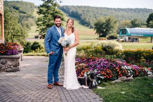 Wedding couple poses on stone patio surrounded by vibrant purple and pink floral gardens at outdoor venue.