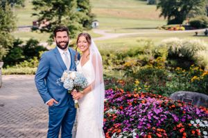 Couple in wedding attire stands on brick path with bright flower garden and rolling hills in background.