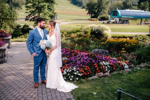 Wedding couple stands on brick walkway surrounded by blooming gardens and mountain scenery.