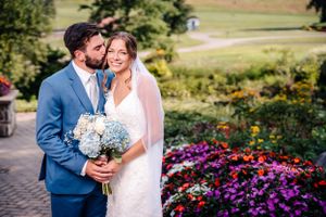 Wedding couple embraces in a garden setting with colorful flower beds and blue hydrangea bouquet.