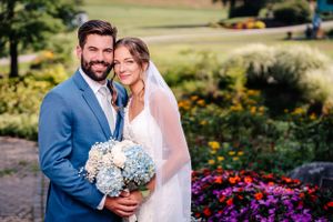 Happy couple poses for wedding portraits in front of a stone wall adorned with purple flowers.