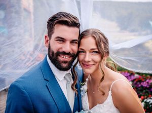 Close-up portrait of newlyweds with veil flowing in breeze against mountain backdrop.