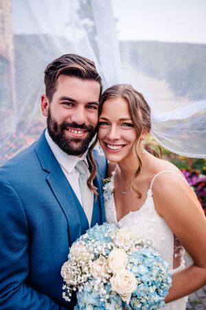 Romantic close-up of wedding couple holding blue and cream floral bouquet against misty background.