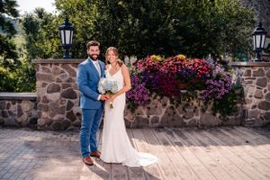 Full-length wedding portrait by stone wall with overflowing flower boxes and classic lanterns.