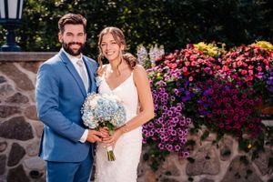 Wedding couple poses by stone wall with vibrant purple petunias and classic outdoor lighting.