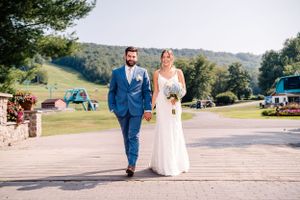 A wedding pair strolls along a paved pathway with green mountains and blue sky in the background.