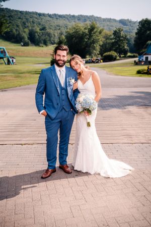 Wedding couple poses on paved pathway with green mountains in background holding light blue bouquet.