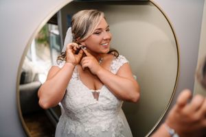Bride putting on jewelry and getting ready in white lace wedding dress reflected in round mirror.