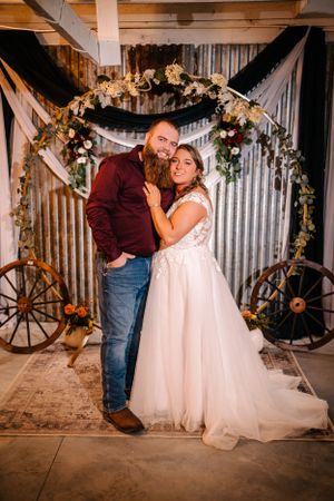 Couple poses under floral arch with wagon wheels and rustic decor at country wedding venue.