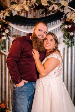 Wedding portrait with couple in burgundy and white against rustic metal wall and floral arch.