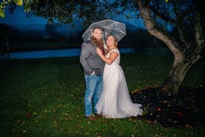 Evening outdoor wedding portrait under clear umbrella with couple on grassy hill.