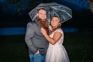 A romantic couple poses under a clear umbrella at dusk in a garden setting.