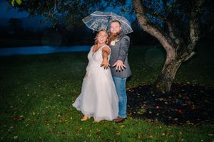 A romantic couple embraces under a clear umbrella on a grassy lawn with a tree.