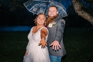 A playful couple poses with outstretched arms under a clear umbrella at night.