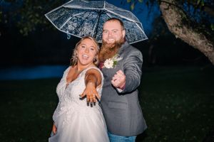 Couple points playfully at camera while sharing a clear umbrella outdoors at dusk.