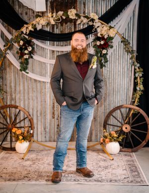 Portrait of person in grey blazer and jeans standing under floral wedding arch with wagon wheels.